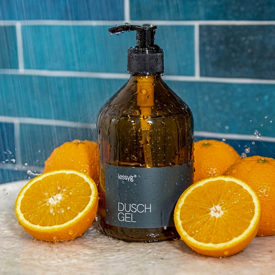 Braune Glas-Pumpflasche mit schwarzem Etikett „Duschgel“ steht auf einem hellen untergrundl in einer modernen Dusche mit Petroleum färbenden Fliesen; daneben liegen halbierte Orangen feine Wassertropfen sind auf Flasche und Glaswand sichtbar.