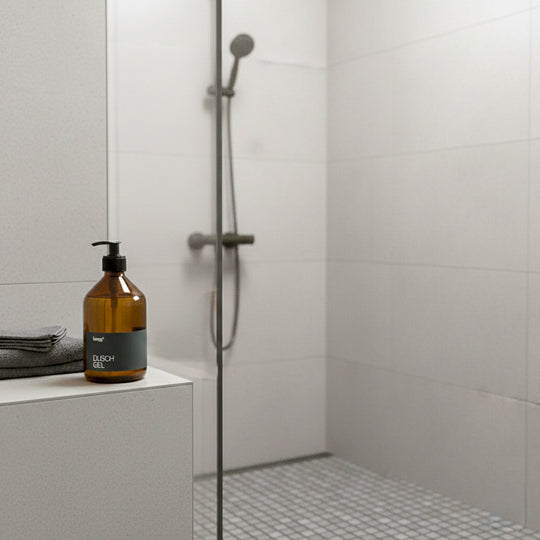 Braune Glas-Pumpflasche mit schwarzem Etikett „Duschgel“ steht auf einem hellen untergrundl in einer modernen Dusche mit weißen Fliesen; daneben liegen Handtücher
